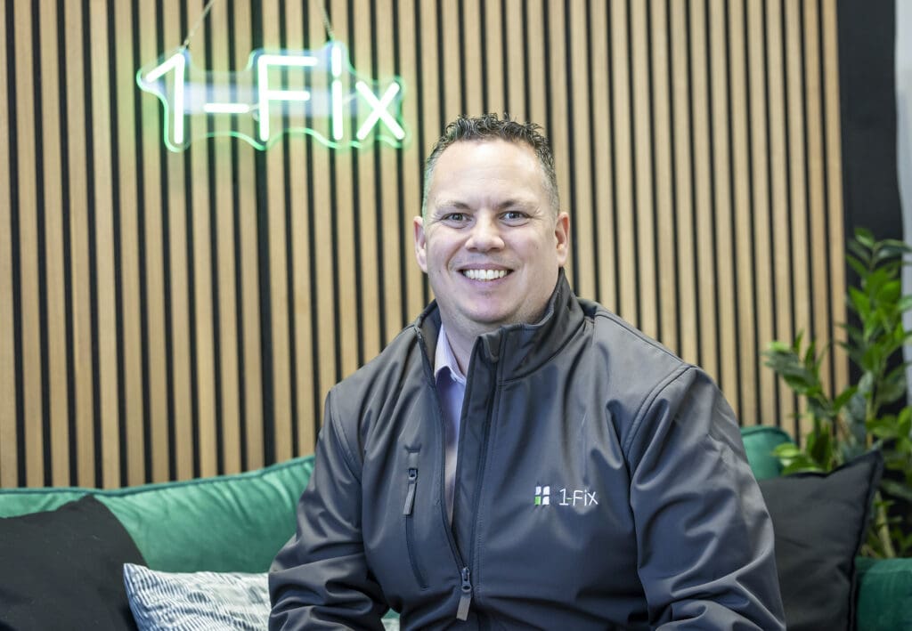 Craig Atkins, Managing Director of 1-Fix is sat on a sofa with the business logo illuminated behind him.